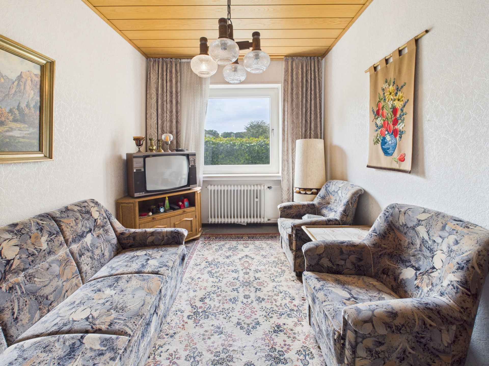 Wohnzimmer mit gemusterten Sofas, Teppich, altem Fernseher und Fenster mit Blick ins Grüne.