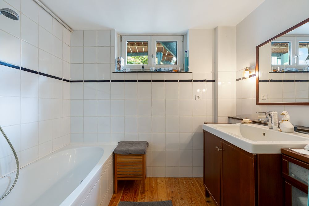Helles Badezimmer mit Badewanne, Fenster, Holzboden und Waschtisch mit Spiegel.