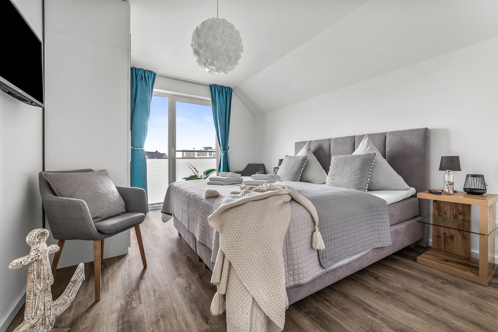 Helles Schlafzimmer mit Doppelbett, grauem Sessel und Balkonzugang mit blauen Vorhängen.