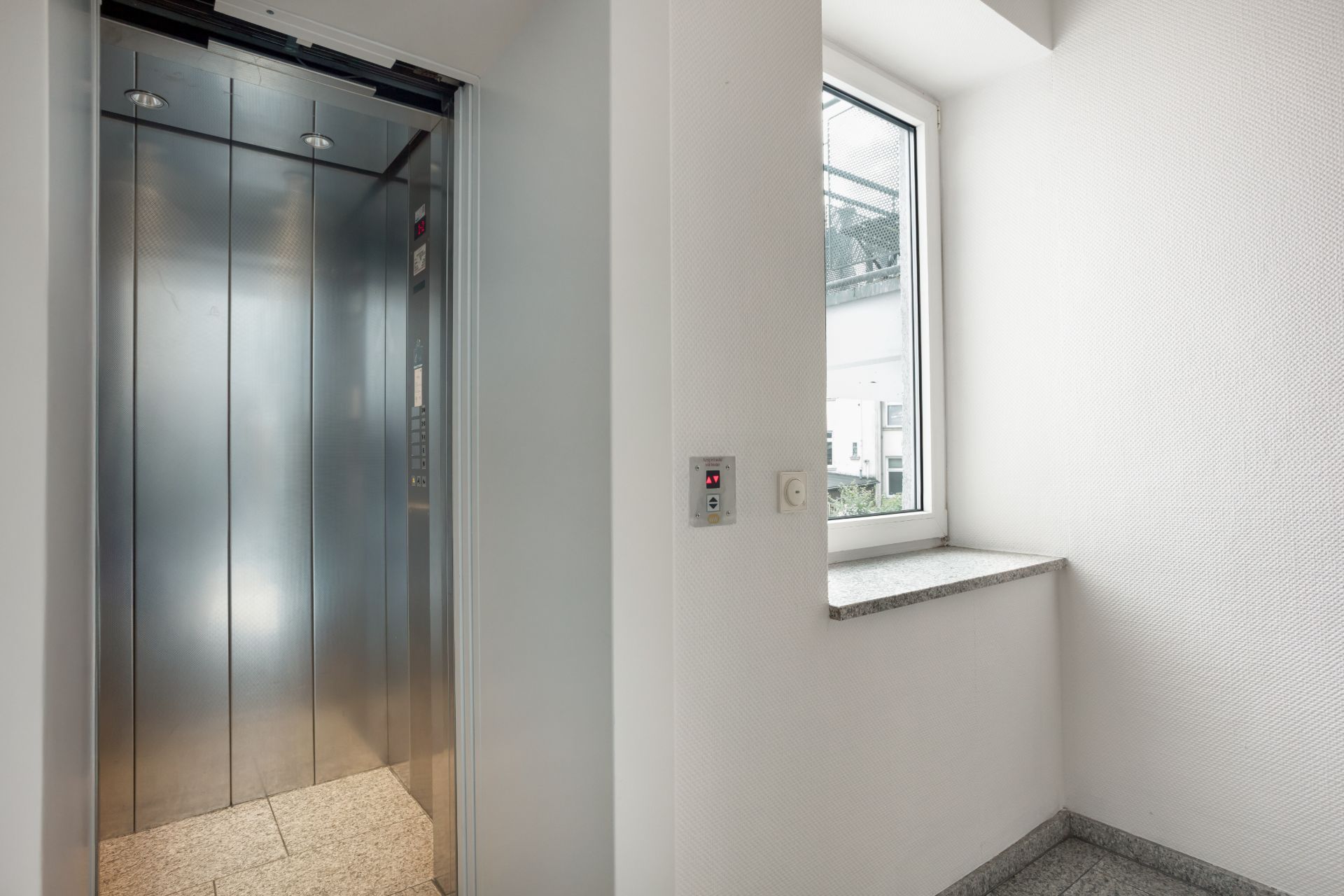 Offener Aufzug im Flur eines Wohngebäudes mit Fenster und grauem Boden.
