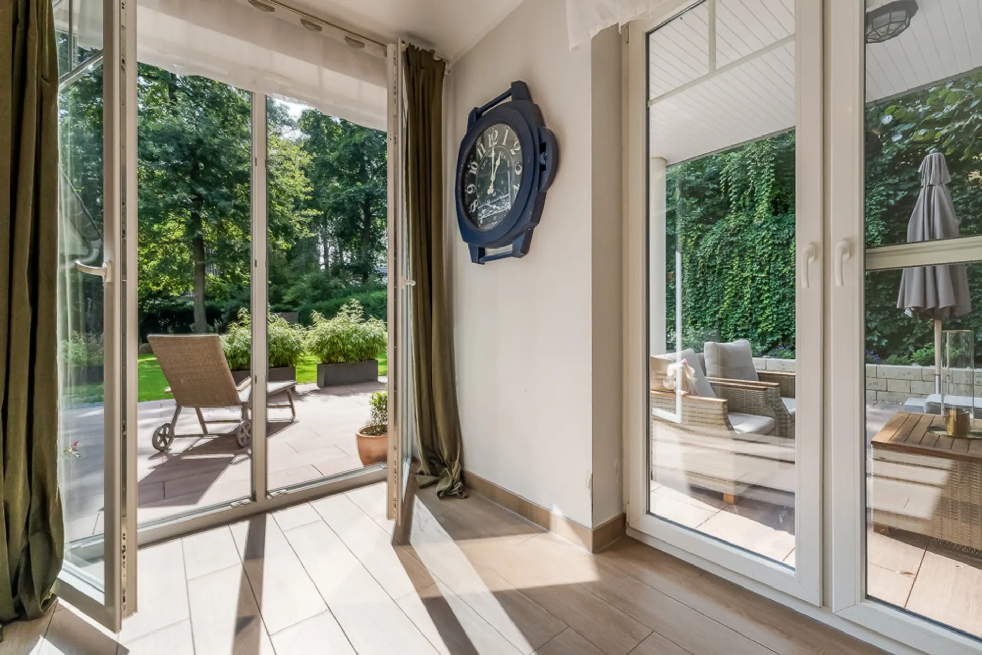 Heller Raum mit großen Glastüren zur Terrasse, Gartenmöbel und grüne Pflanzen draußen sichtbar.