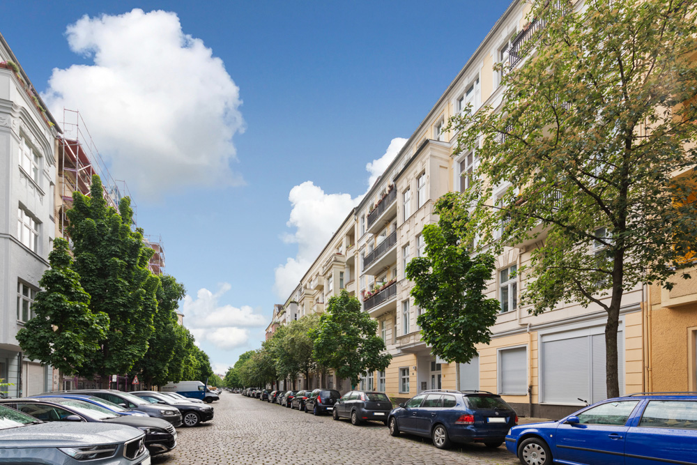 Baumbestandene Wohnstraße mit parkenden Autos und mehrstöckigen Altbauten bei blauem Himmel.