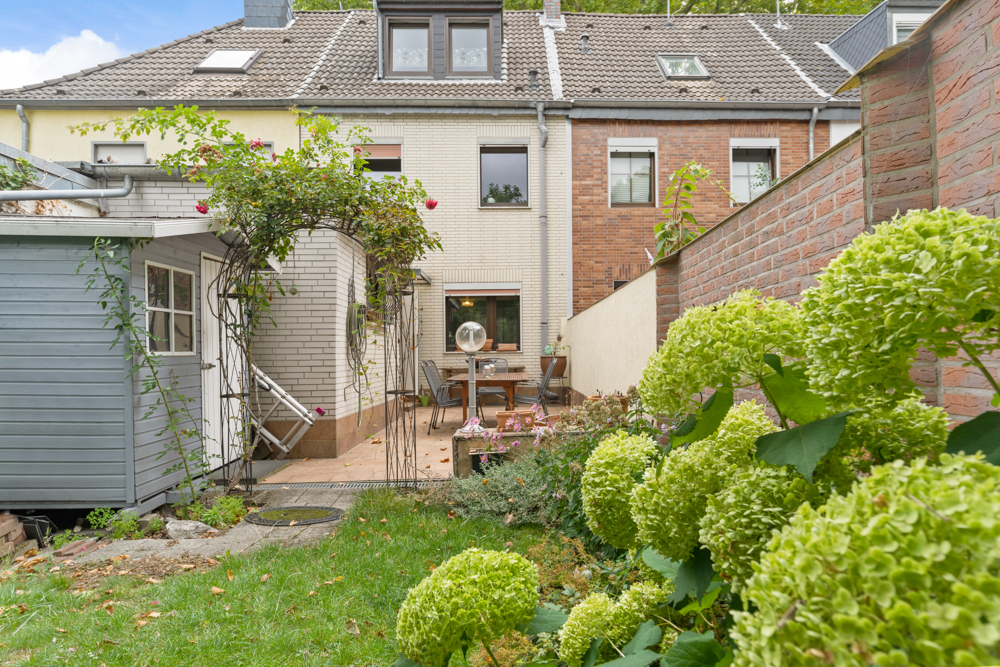 Garten mit Terrasse, Sitzgruppe, Blumen und Gartenhaus hinter einem Wohnhaus.
