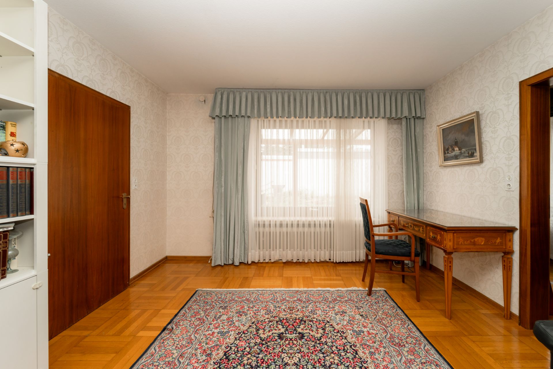 Helles Zimmer mit Holzboden, Teppich, Schreibtisch, Stuhl und großem Fenster mit Vorhängen.