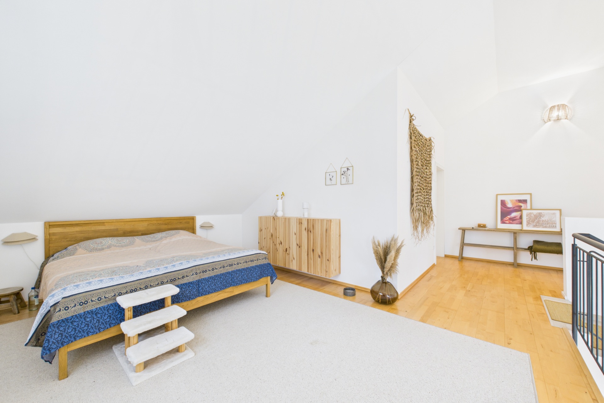 Helles Schlafzimmer mit Holzfußboden, Doppelbett, Kommode und dekorativem Teppich an der Wand.