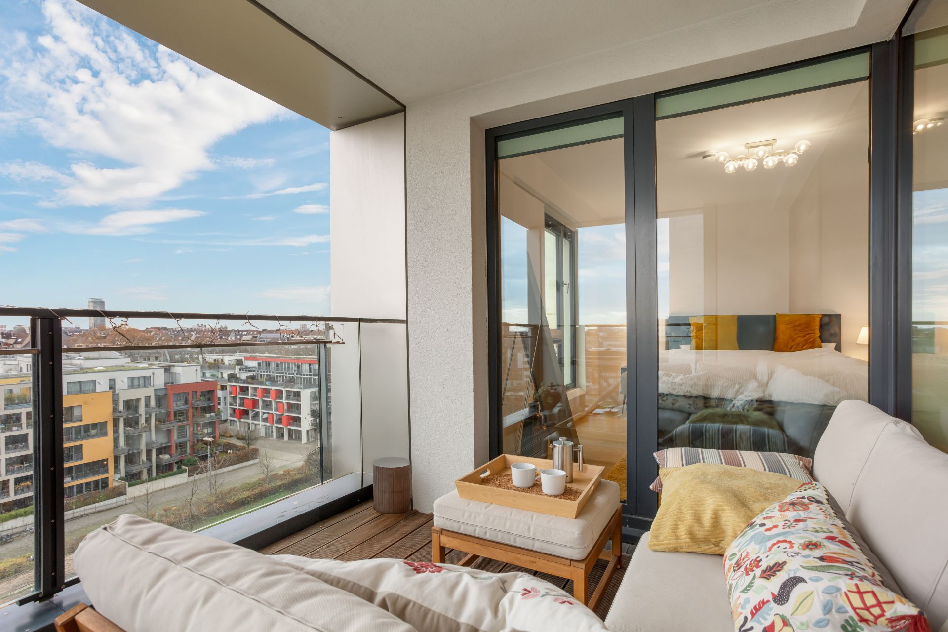 Balkon mit Sofa, Tisch und Blick auf Wohngebäude, Schlafzimmer durch Glastür sichtbar.
