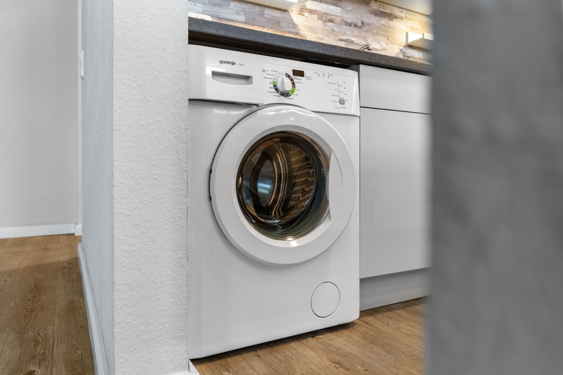 Eine weiße Waschmaschine steht unter einer Küchenarbeitsplatte auf Holzboden.