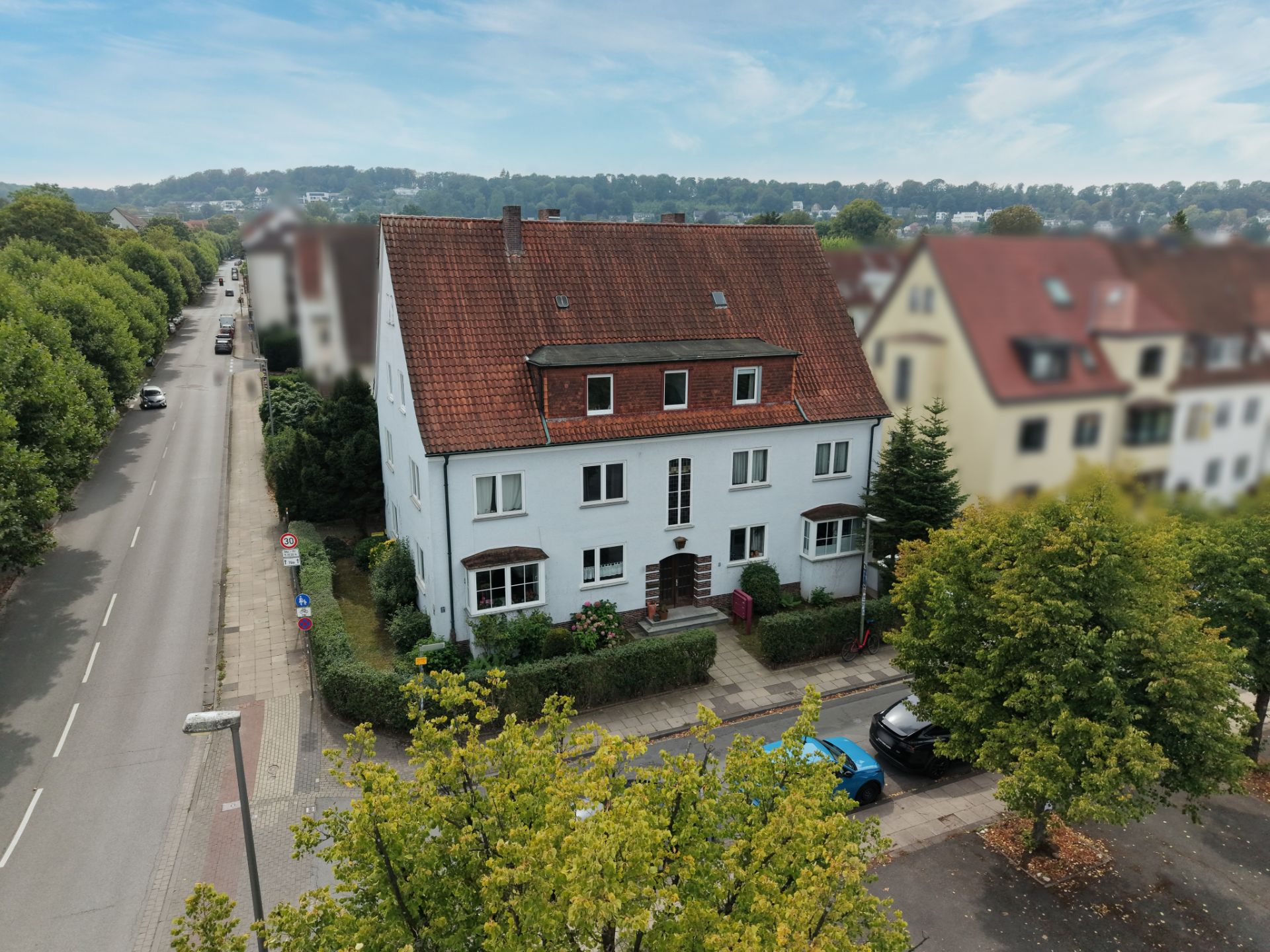 Mehrfamilienhaus mit rotem Dach an einer von Bäumen gesäumten Straße, Autos parken davor.