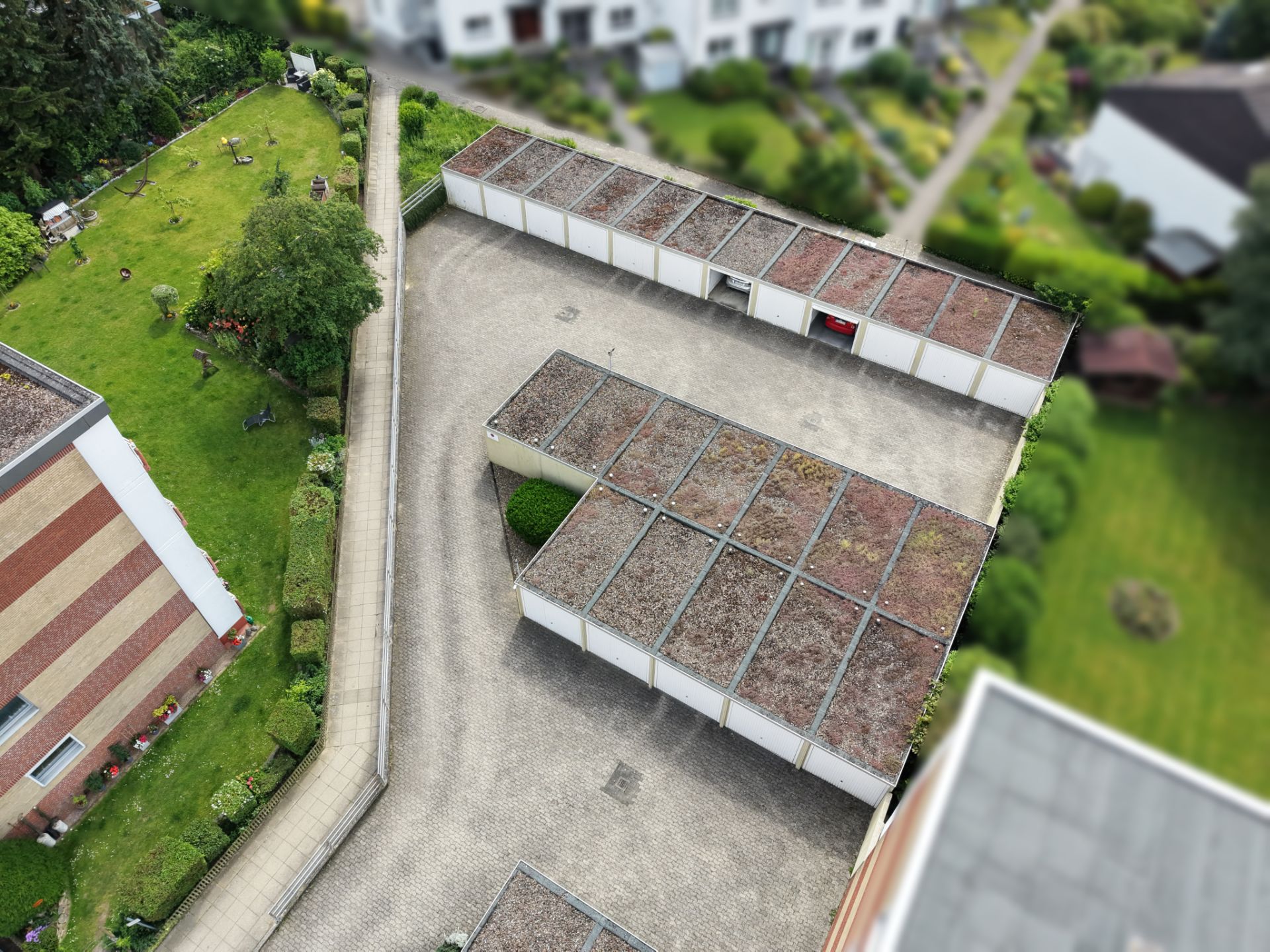 Mehrere Garagen mit Flachdach stehen auf einem gepflasterten Hof neben einem Wohngebäude und Garten.
