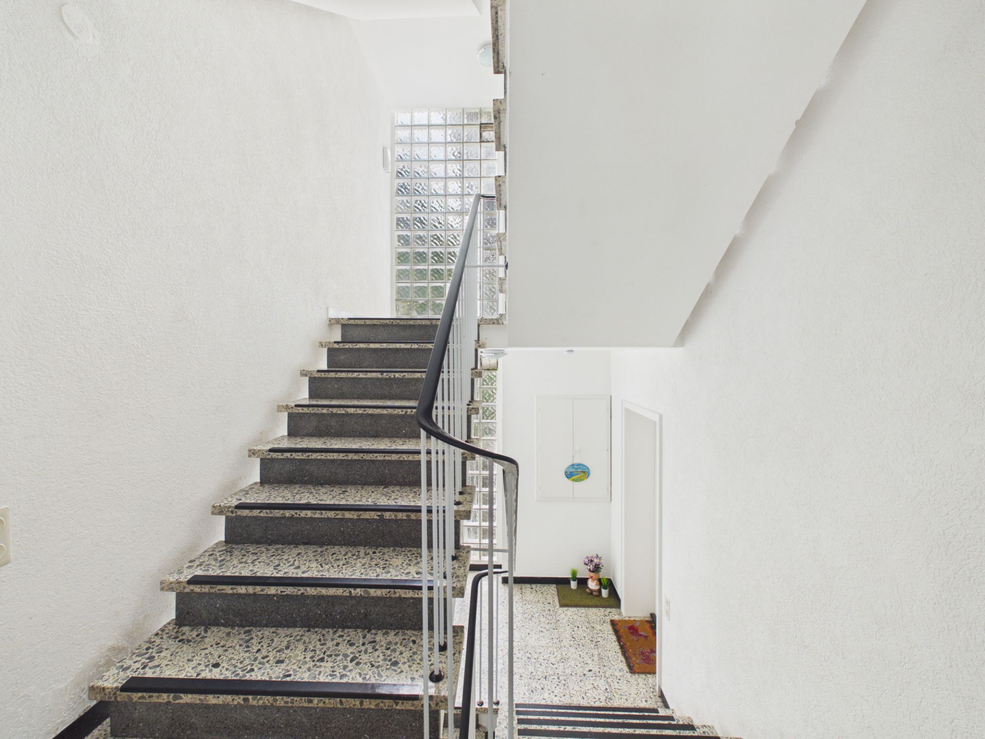 Treppenhaus mit grauer Steintreppe, weißer Wand und Glasbausteinfenster im Wohngebäude.