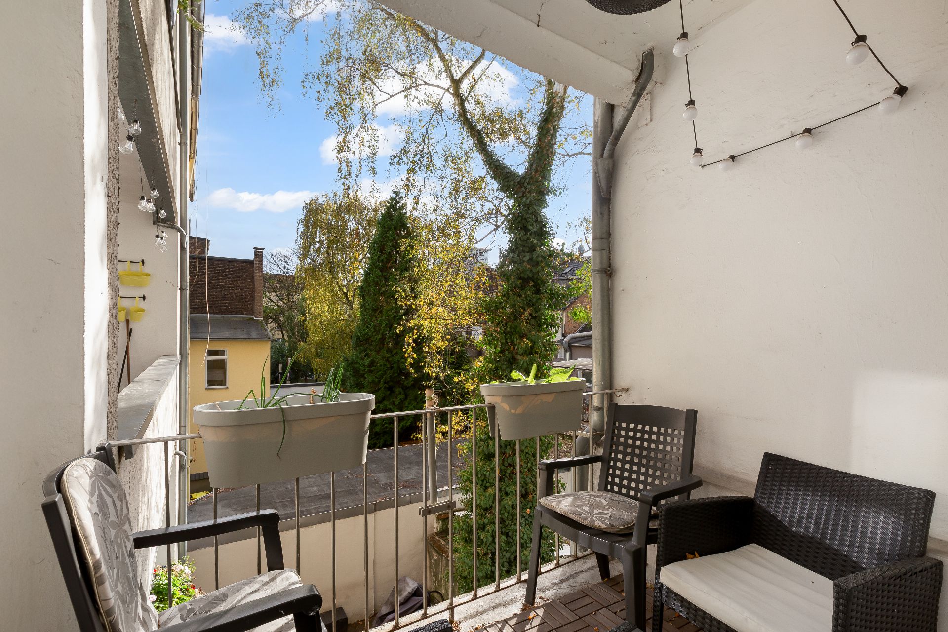 Kleiner Balkon mit zwei Stühlen, einem Sitzbank, Blumentöpfen am Geländer und Blick auf Bäume.