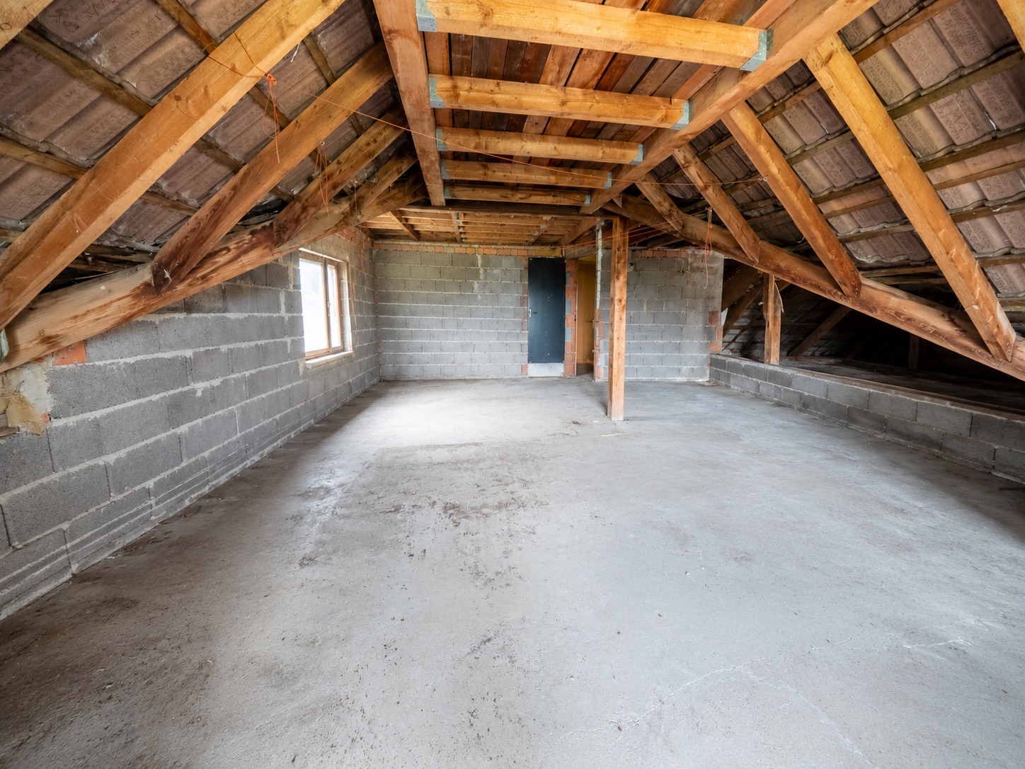 Leerstehender Dachboden mit Sicht auf Holzbalken, Betonboden und einem kleinen Fenster.