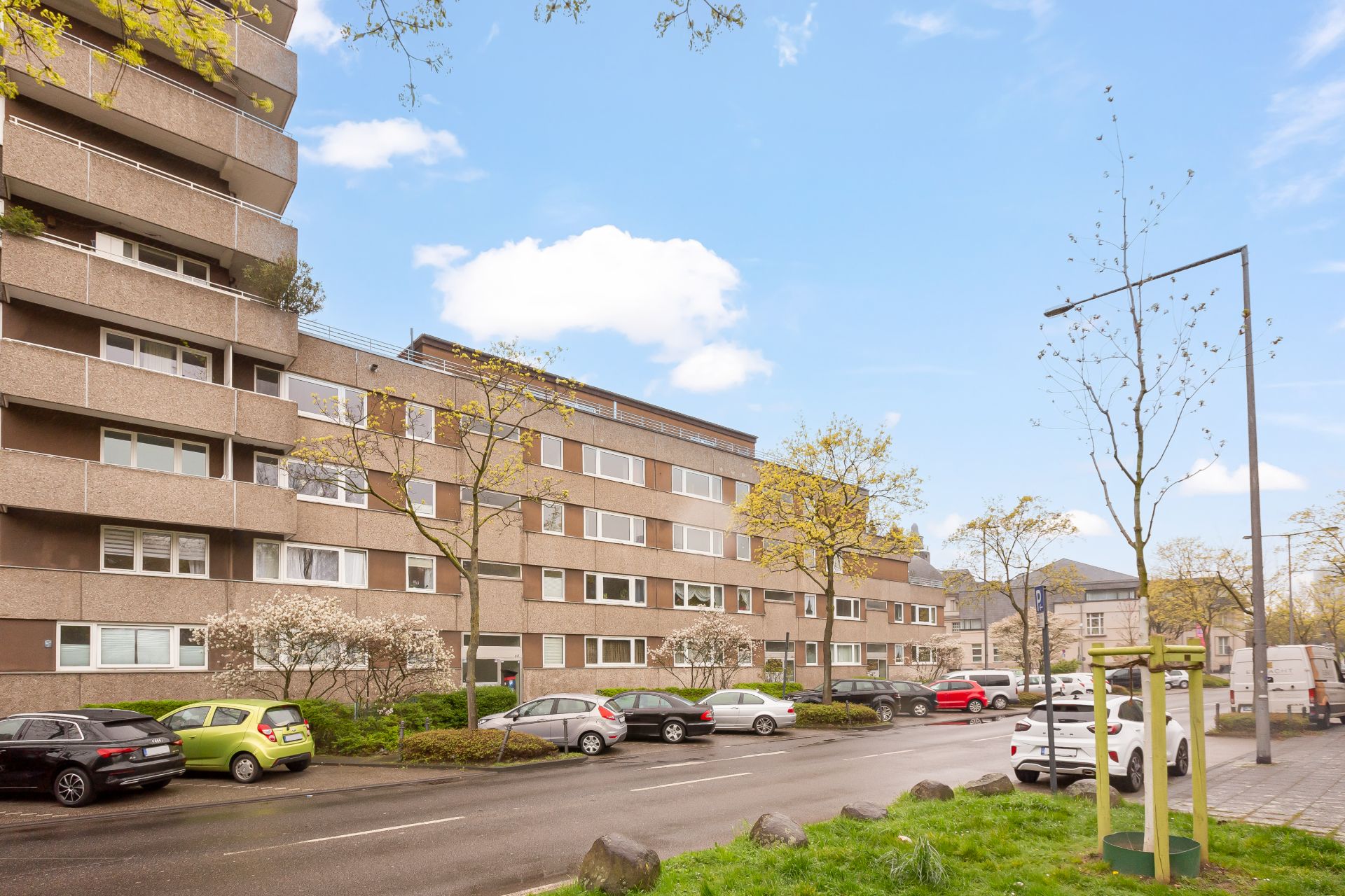 Mehrstöckiges Wohnhaus mit parkenden Autos und Bäumen an einer Straße bei Tageslicht.