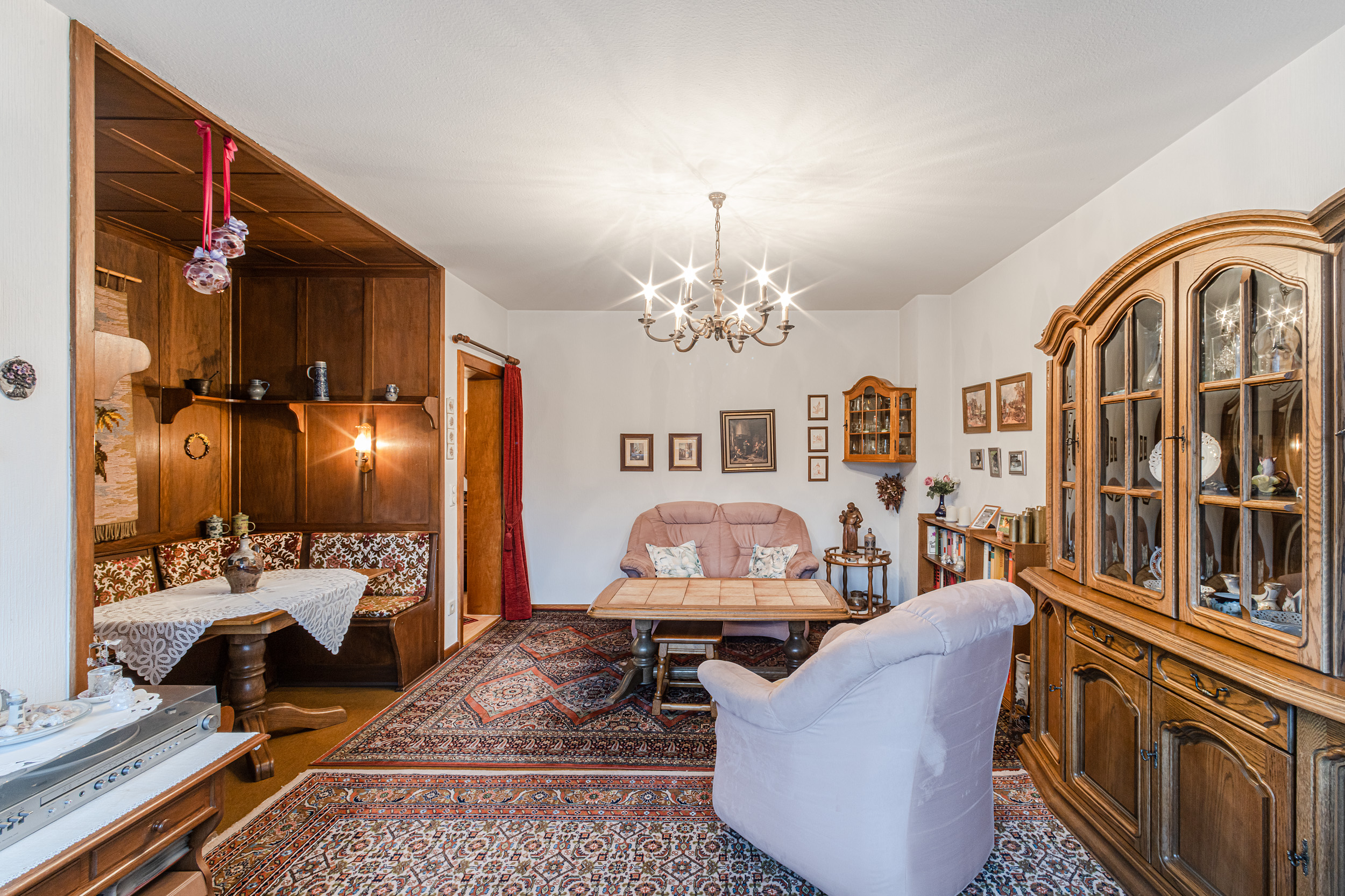 Gemütliches Wohnzimmer mit Teppich, Sofa, Sessel, Esstisch, Vitrine und Eckbank mit Holzwand.