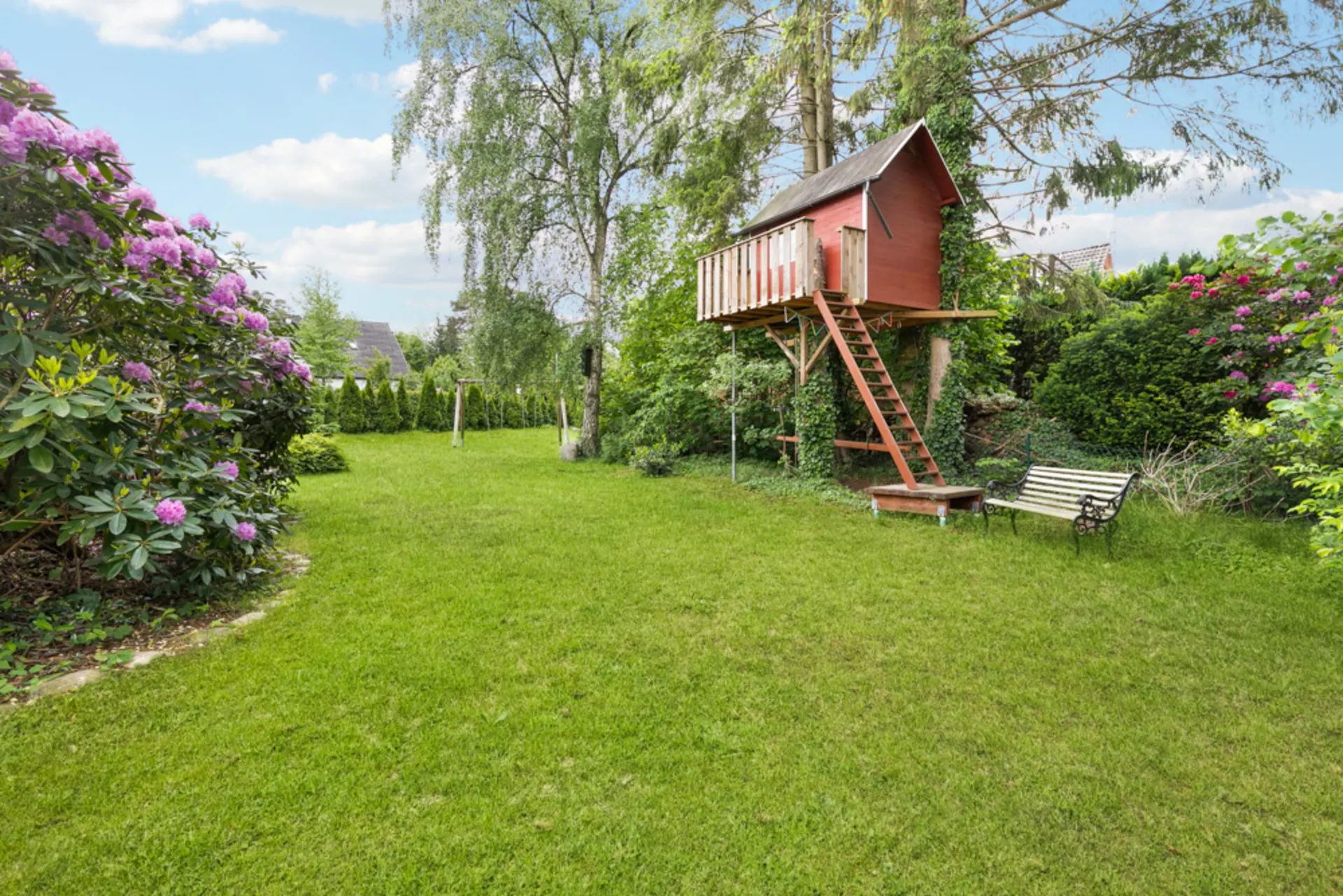 Garten mit grünem Rasen, Baumhaus auf Stelzen, Blütensträuchern und einer Gartenbank.
