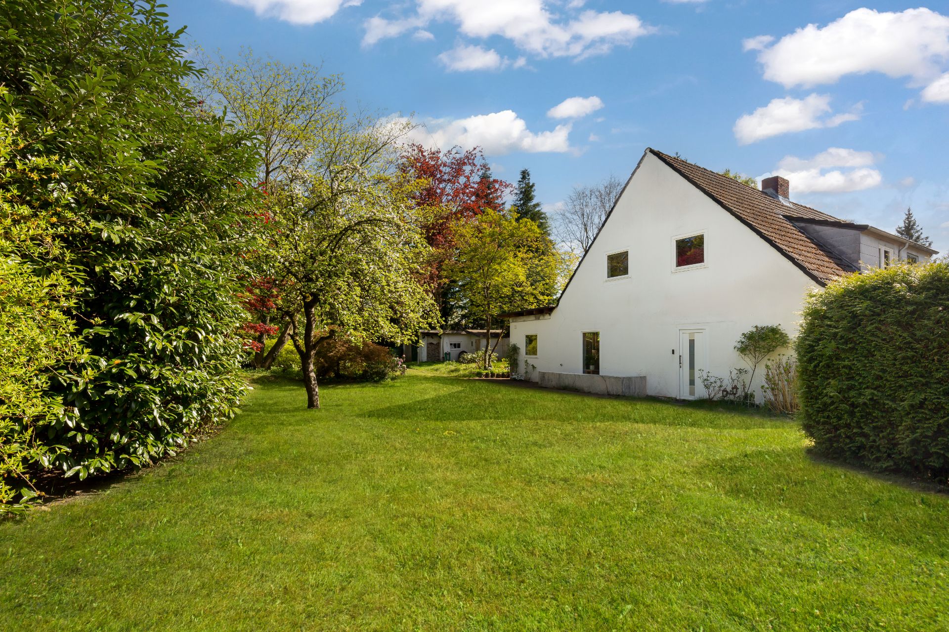Haus mit weißer Fassade und großem Garten mit Bäumen unter blauem Himmel.