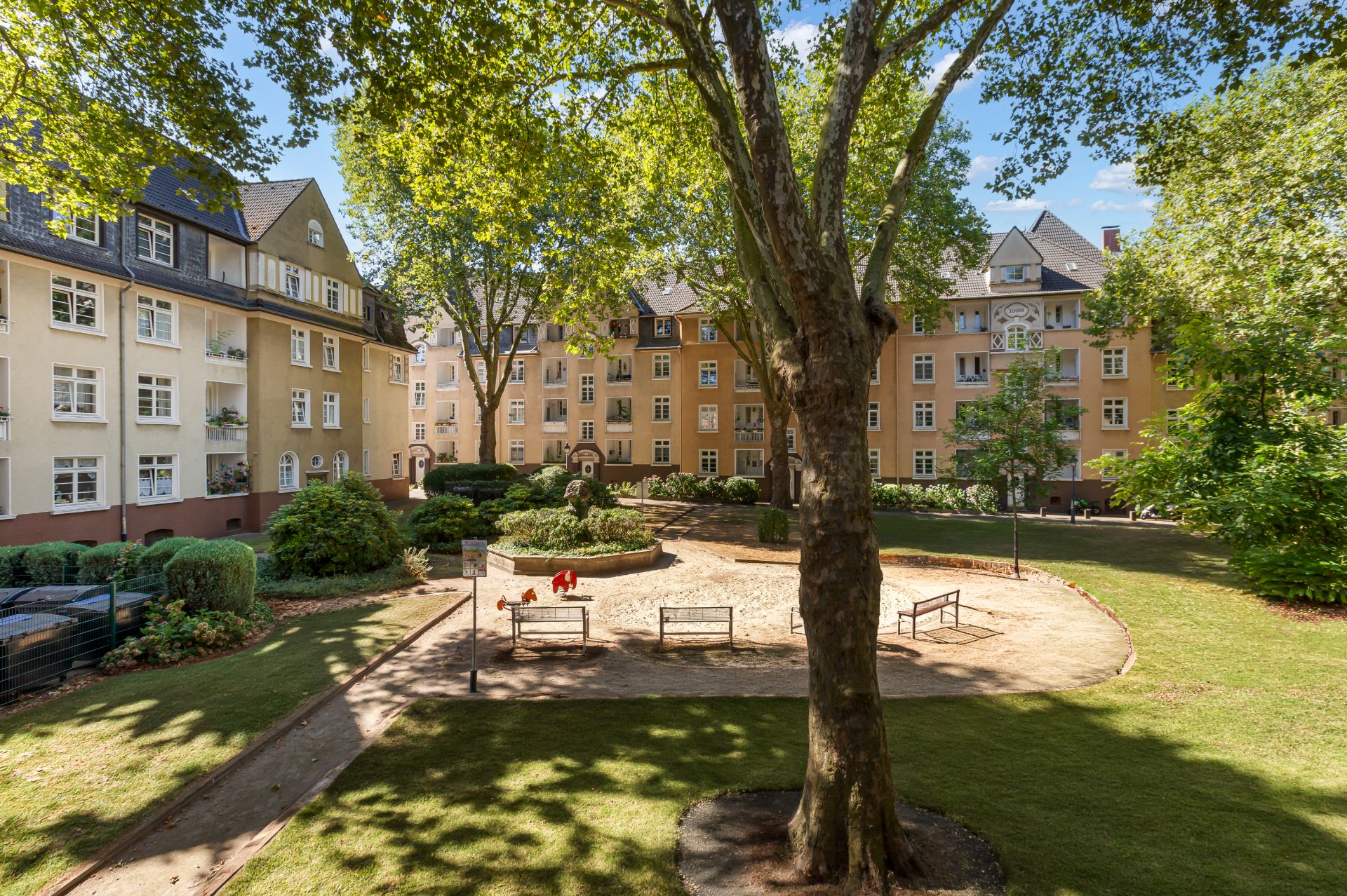 Wohnhof mit Sandspielplatz, Bänken, Bäumen und umliegenden Mehrfamilienhäusern bei Sonnenschein.