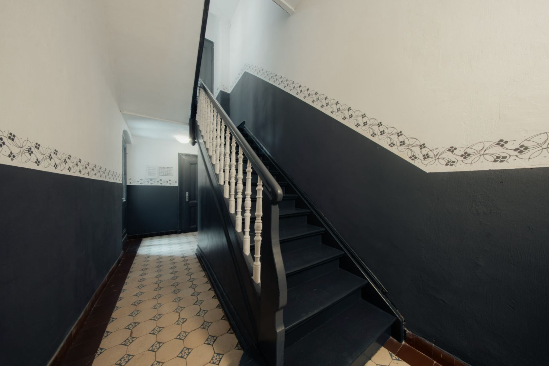 Treppe mit dunklem Geländer und gemusterten Fliesen in einem Wohnhausflur.
