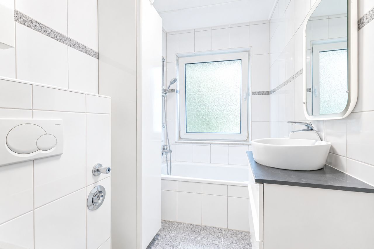 Helles Badezimmer mit Badewanne, Waschbecken, Spiegel und Fenster, weiße Fliesen an Wänden und Boden.