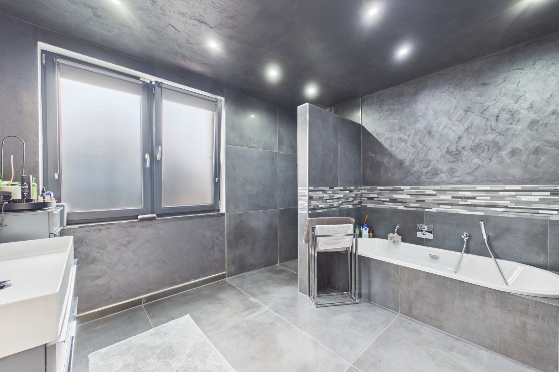 Modernes Bad mit grauen Fliesen, Badewanne, Waschbecken und großem Fenster mit Milchglas.