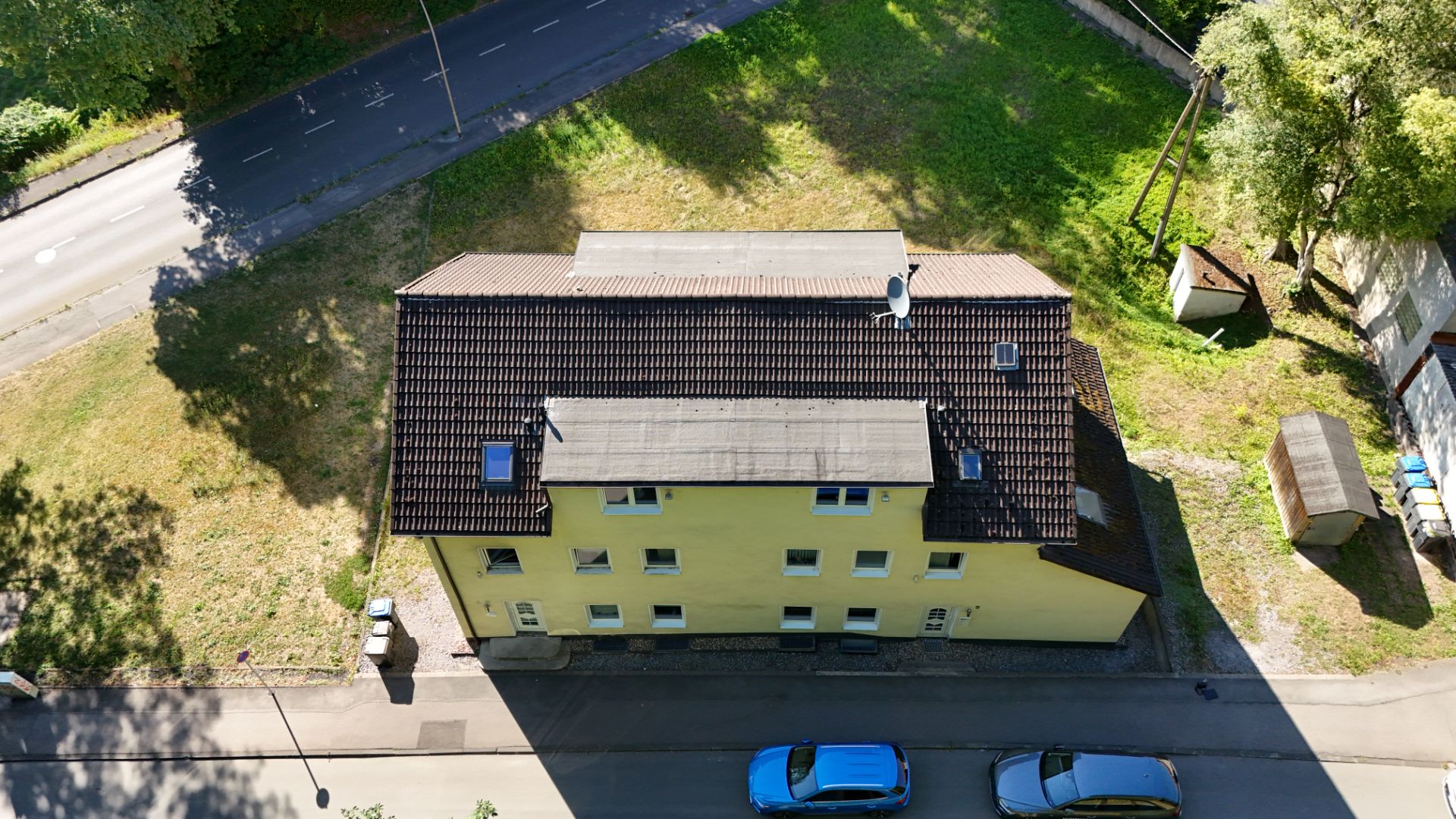 Luftaufnahme eines gelben Wohngebäudes mit braunem Dach, geparkten Autos und umliegendem Grün.