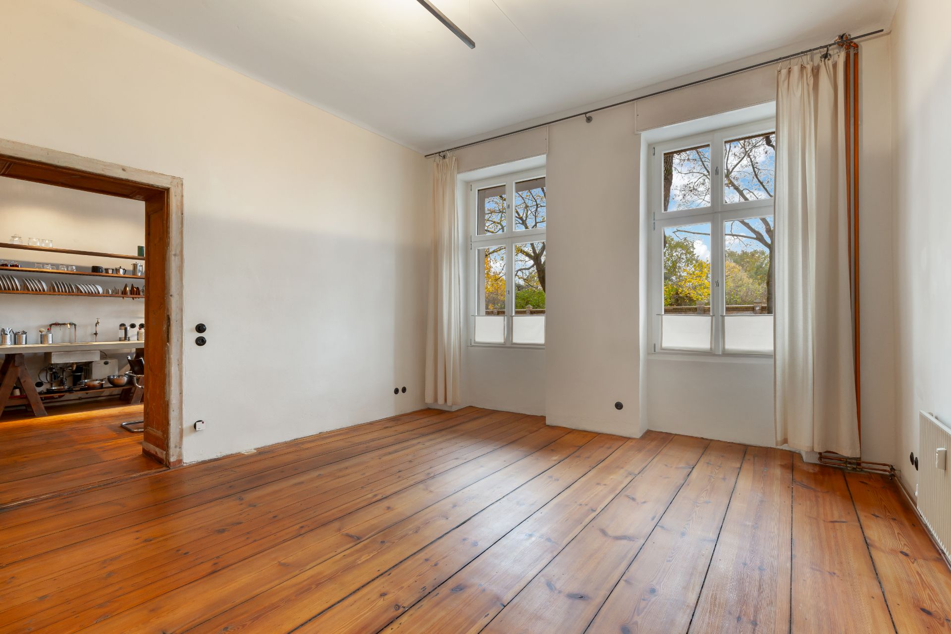 Helles Zimmer mit Holzboden, zwei großen Fenstern und Blick in eine angrenzende Küche.