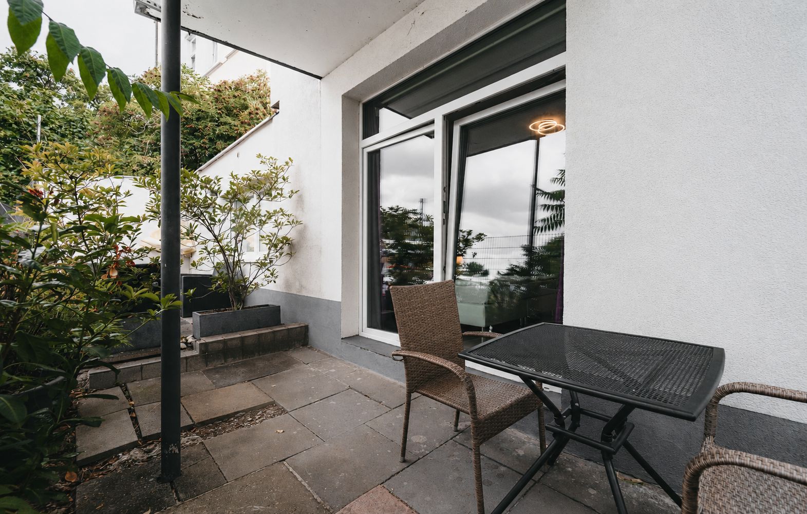 Kleine Terrasse mit zwei Stühlen, Tisch und Pflanzen neben einem Fenster.