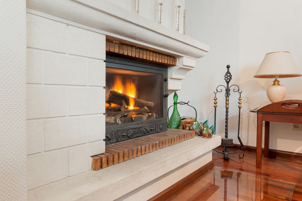 Kamin mit brennendem Feuer, daneben ein Tisch mit Lampe und Kaminbesteck auf Holzboden.