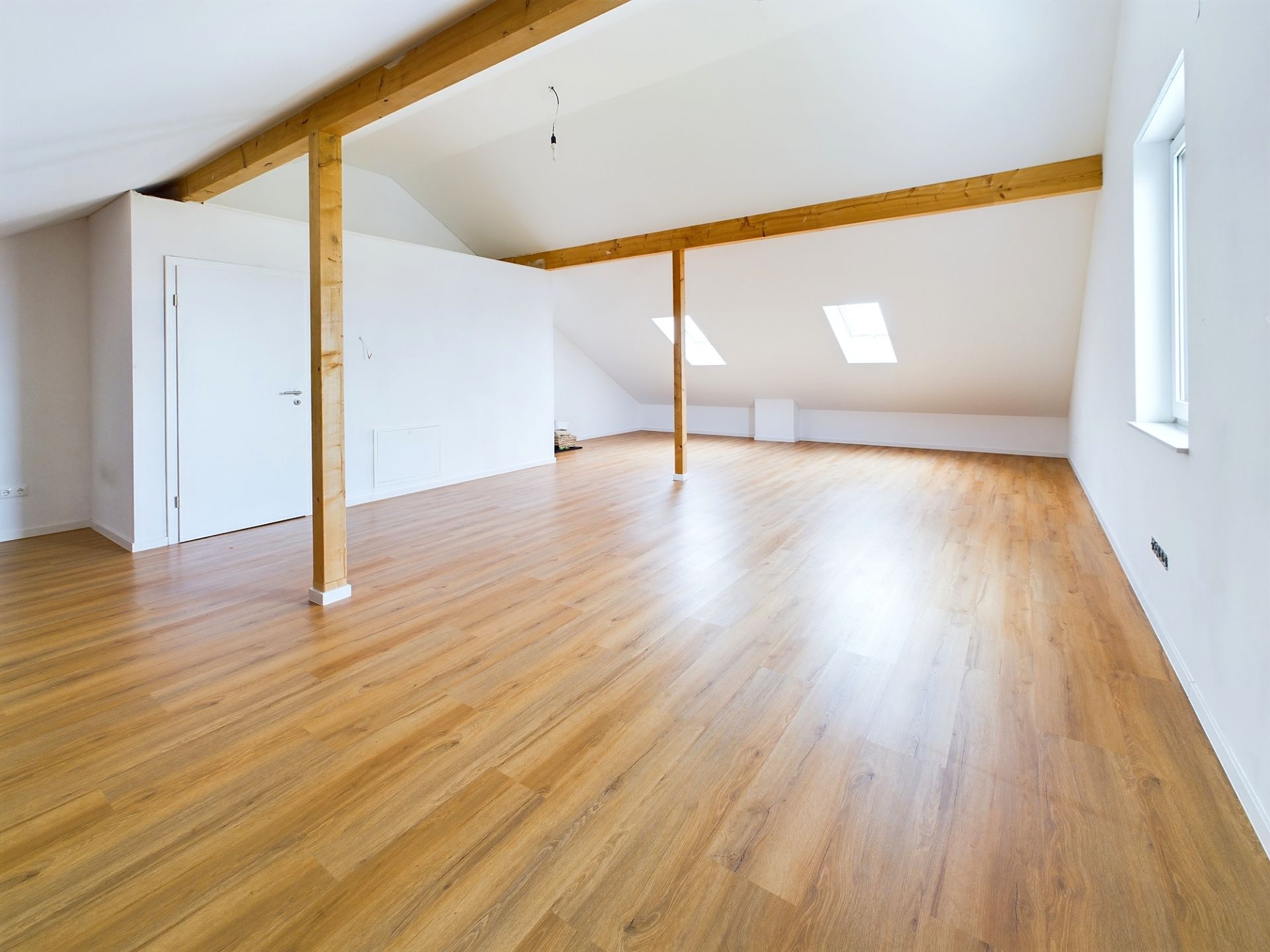 Heller Dachgeschossraum mit Holzboden, Balken und Fenster im Dach.