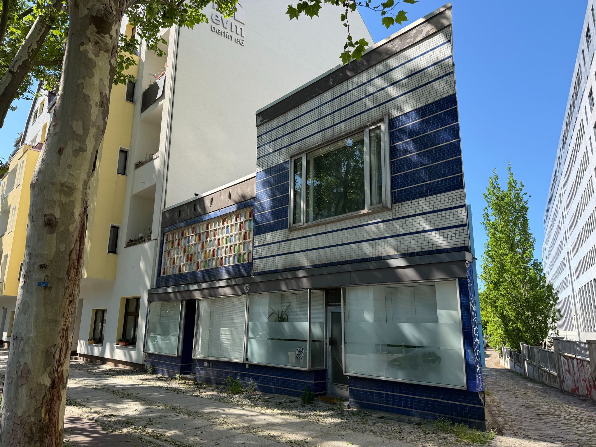Zweistöckiges Gebäude mit blauer Fassade und großen Fenstern, daneben ein Baum.