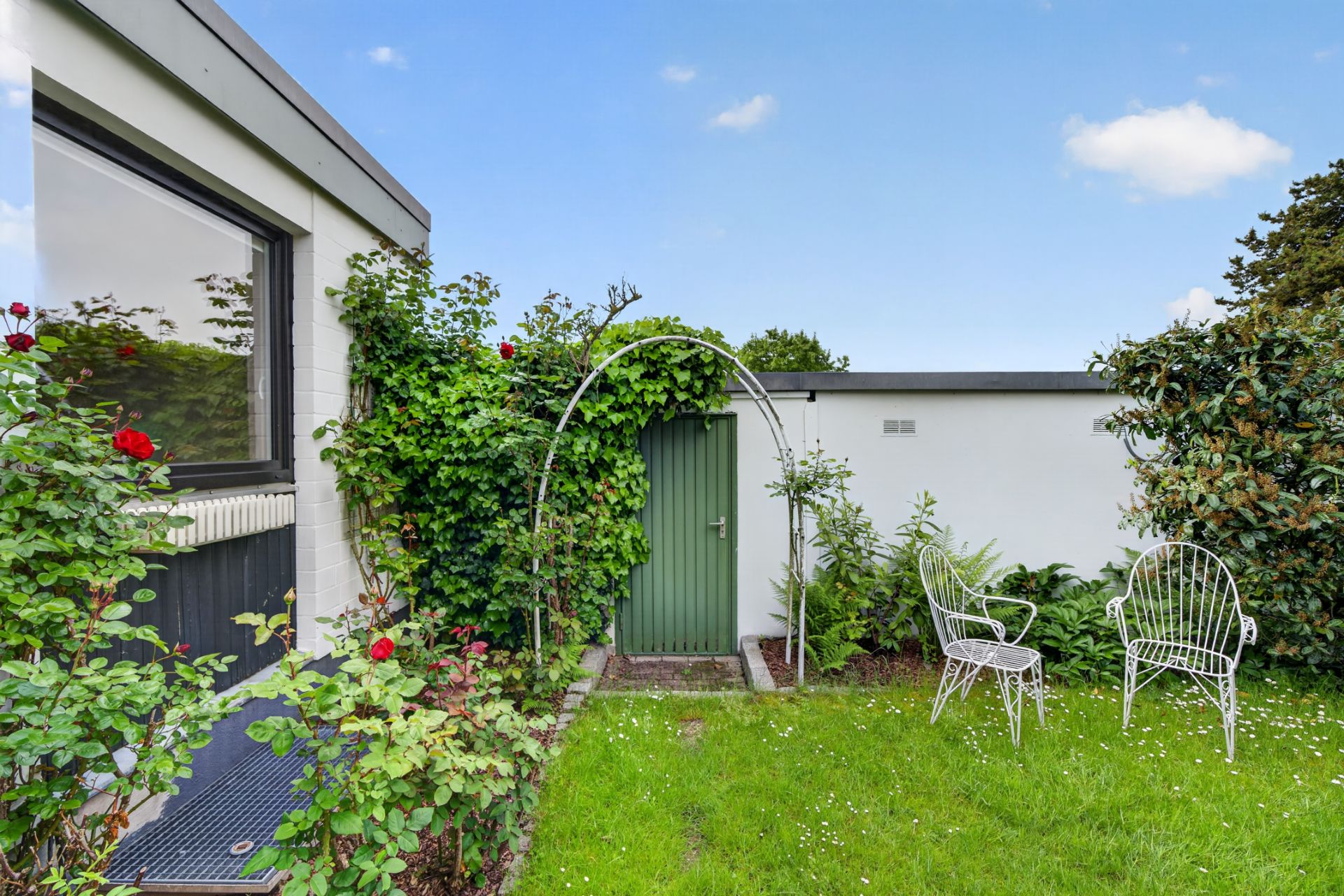 Garten mit grünem Tor, Rosen, Rankbogen und zwei weißen Metallstühlen auf Rasen.