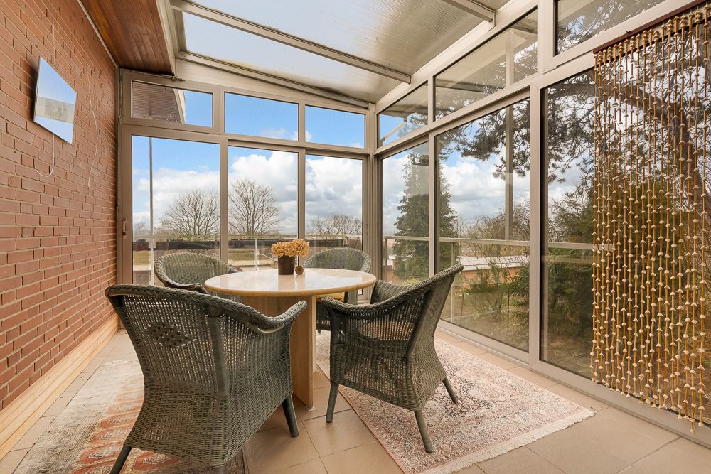 Veranda mit Glaswänden, Rundtisch und vier Korbstühlen, Blick auf Bäume draußen.