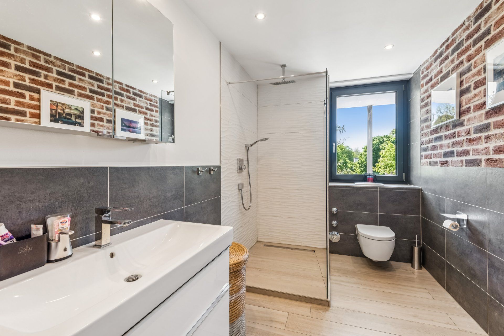 Modernes Badezimmer mit Dusche, Waschtisch, WC und Backsteinwand, großes Fenster im Hintergrund.