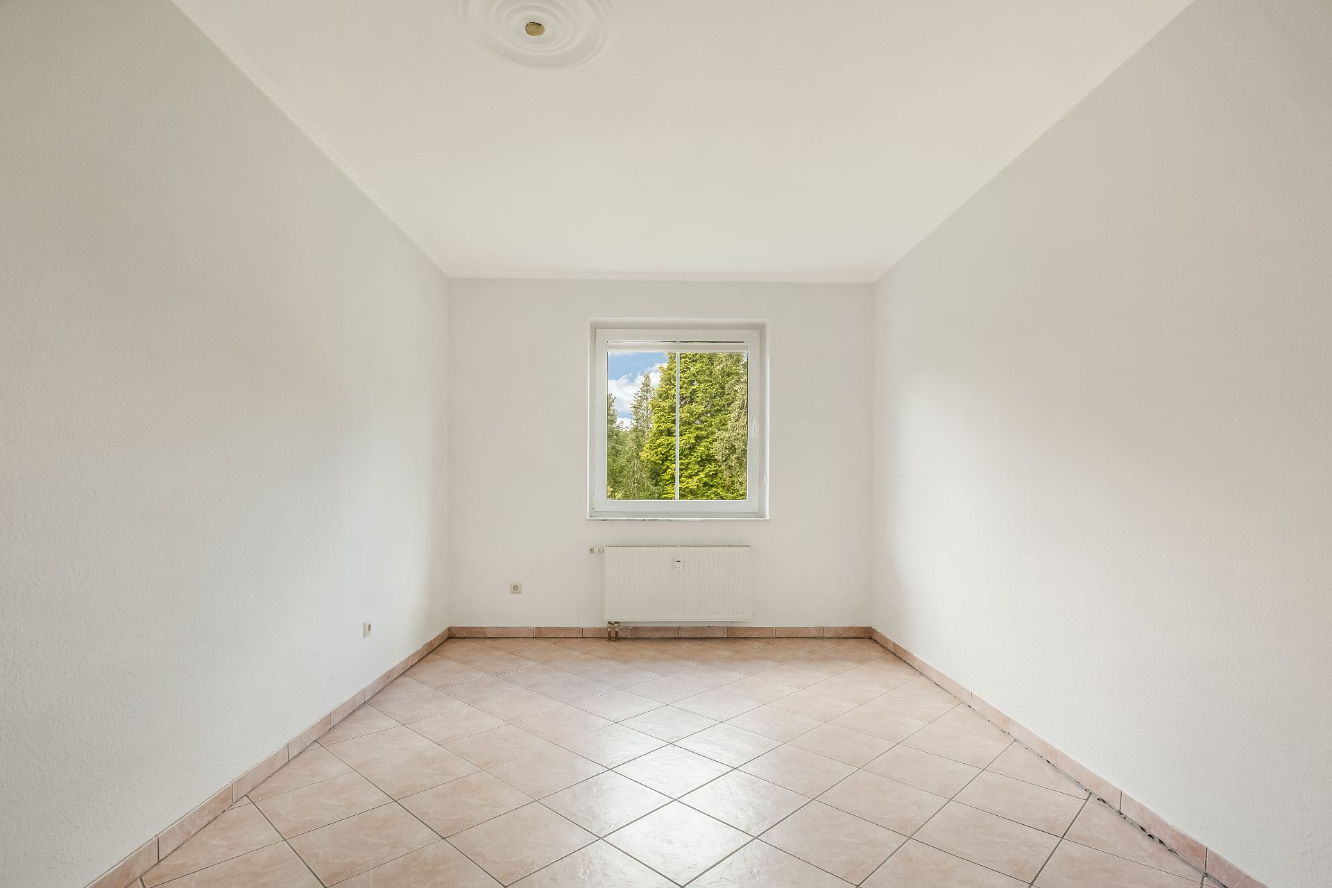 Leerer, heller Raum mit Fliesenboden und einem Fenster mit Blick auf Bäume.