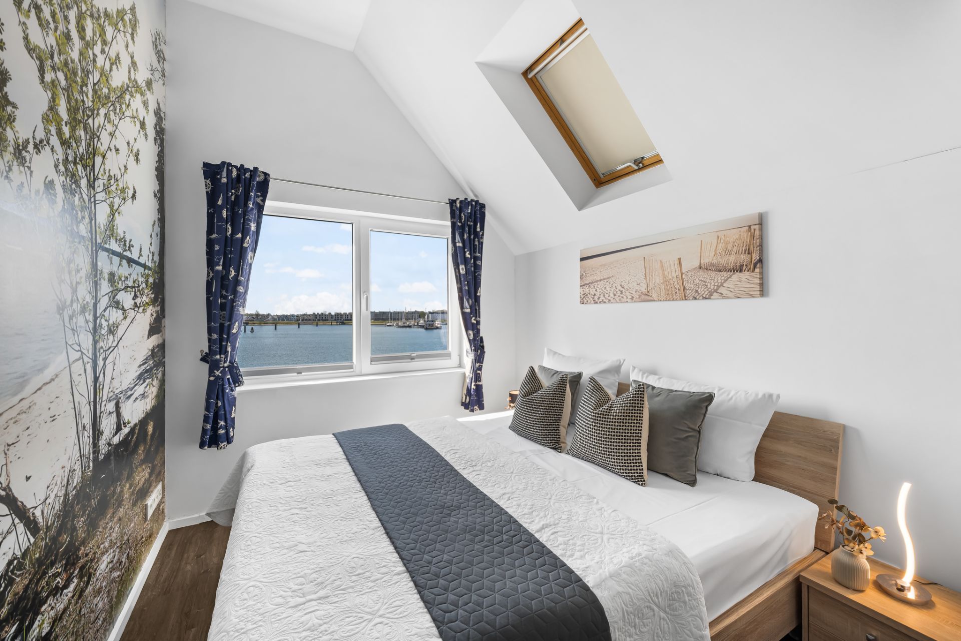 Helles Schlafzimmer mit Doppelbett, Fenster mit Vorhängen und Blick auf das Wasser, Dachfenster und Wandbild.