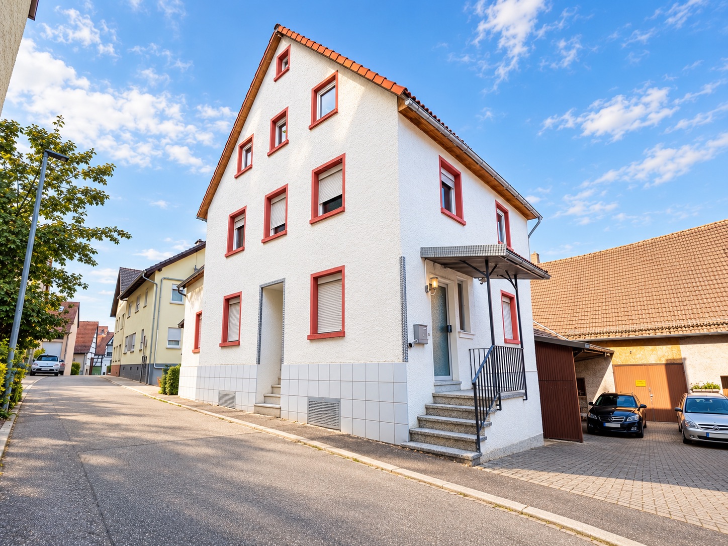Weißes Wohnhaus mit rotem Dach und mehreren Fenstern an einer ruhigen Straße.