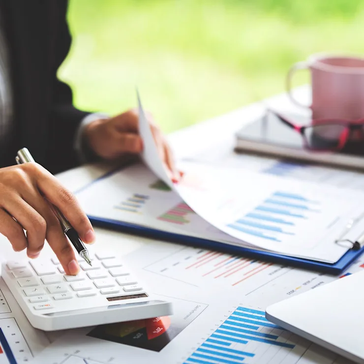 Person arbeitet mit Taschenrechner, Stift und Diagrammen an einem Schreibtisch.
