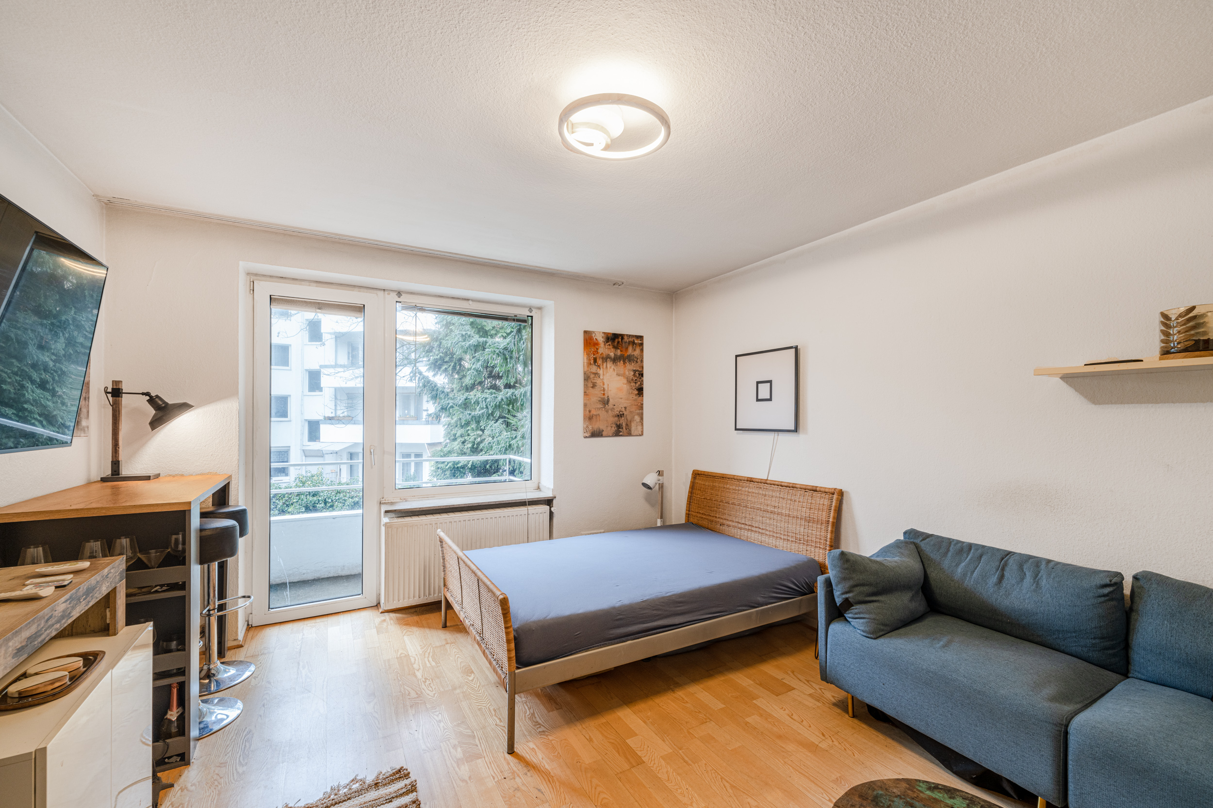 Wohnzimmer mit Bett, Sofa, Schreibtisch und Zugang zum Balkon, helle Holzfußböden.