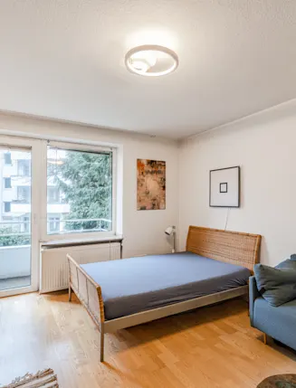 Wohnzimmer mit Bett, Sofa, Schreibtisch und Zugang zum Balkon, helle Holzfußböden.