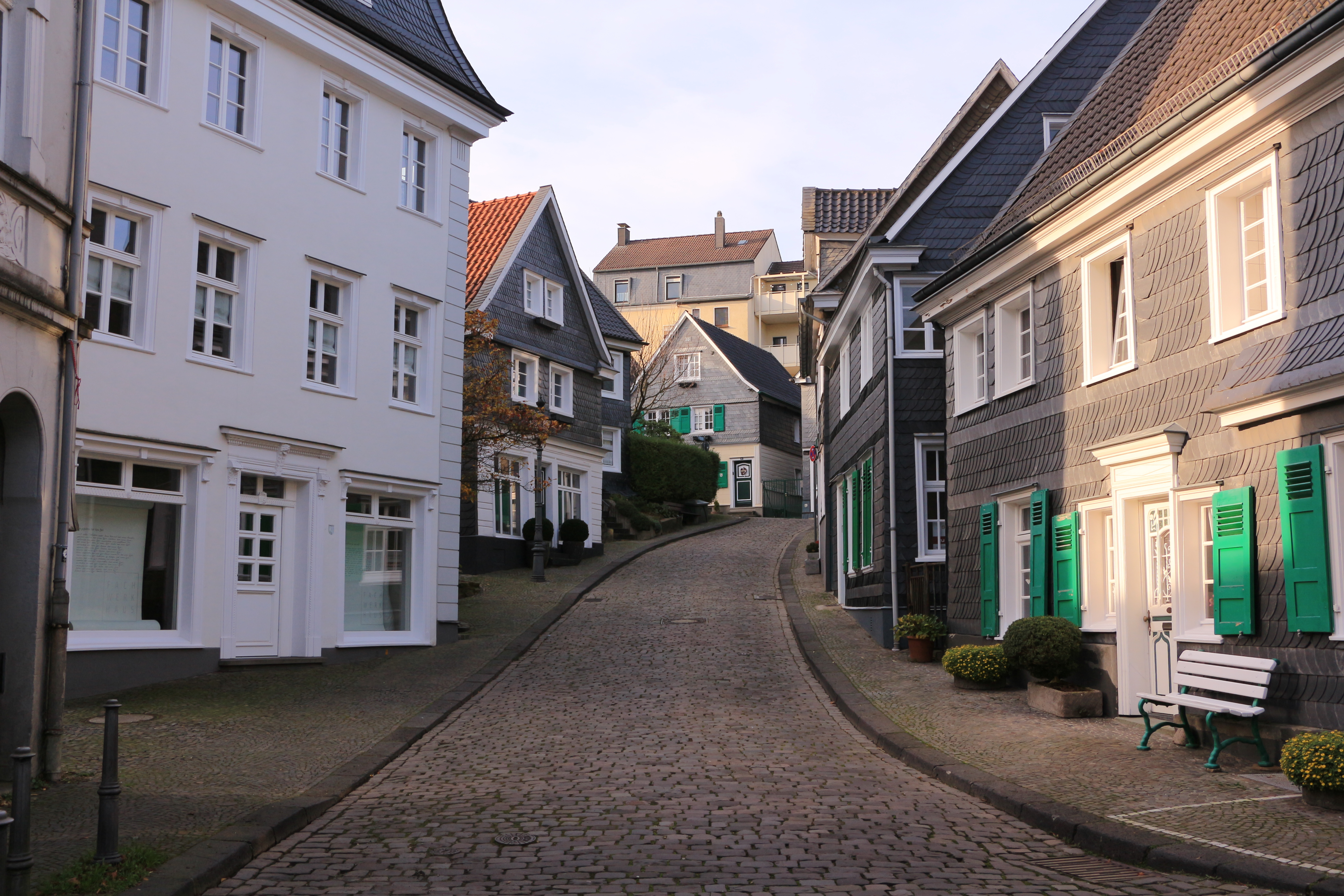 Gepflasterte Straße mit Wohnhäusern, weißen und grauen Fassaden, grünen Fensterläden.
