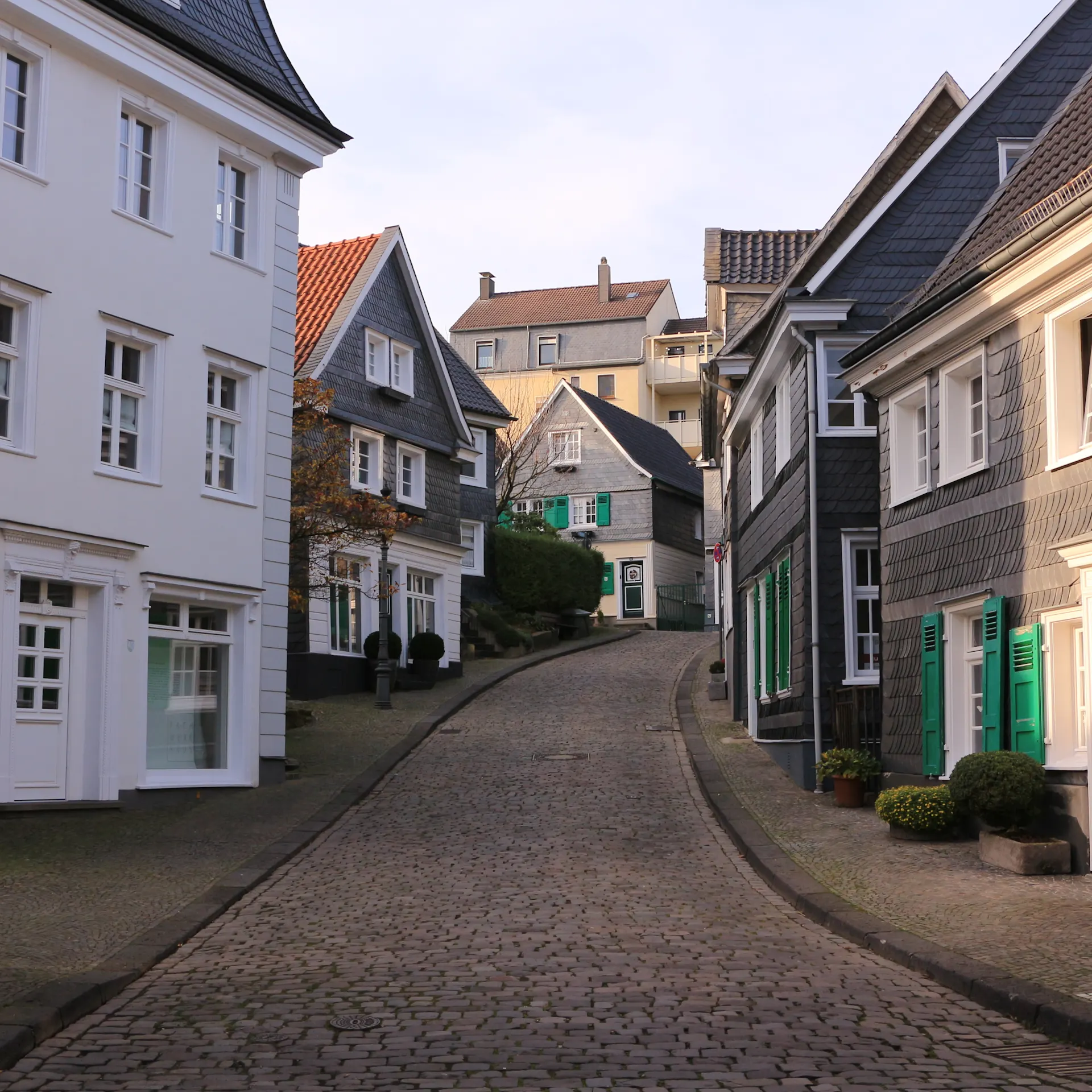 Gepflasterte Straße mit Wohnhäusern, weißen und grauen Fassaden, grünen Fensterläden.