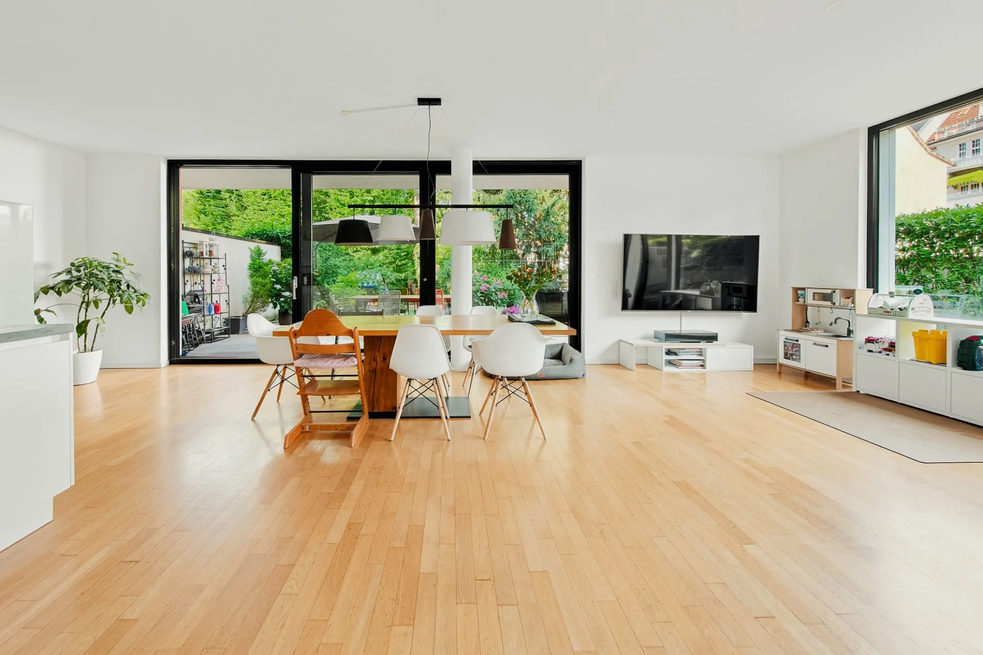 Helles Wohnzimmer mit Holzboden, Esstisch, Stühlen, TV an der Wand und großen Fenstern zum Garten.