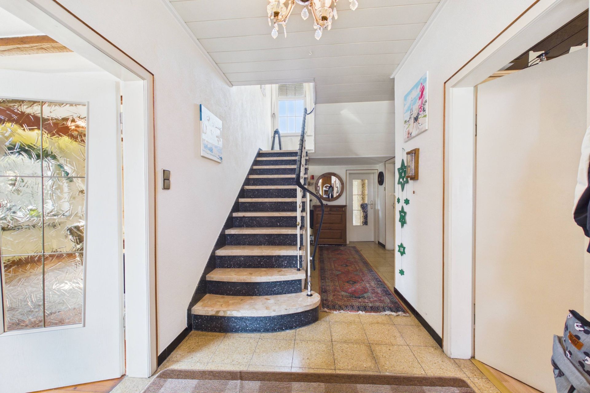 Heller Flur mit Treppe, Kronleuchter und mehreren offenen Türen zu angrenzenden Räumen.