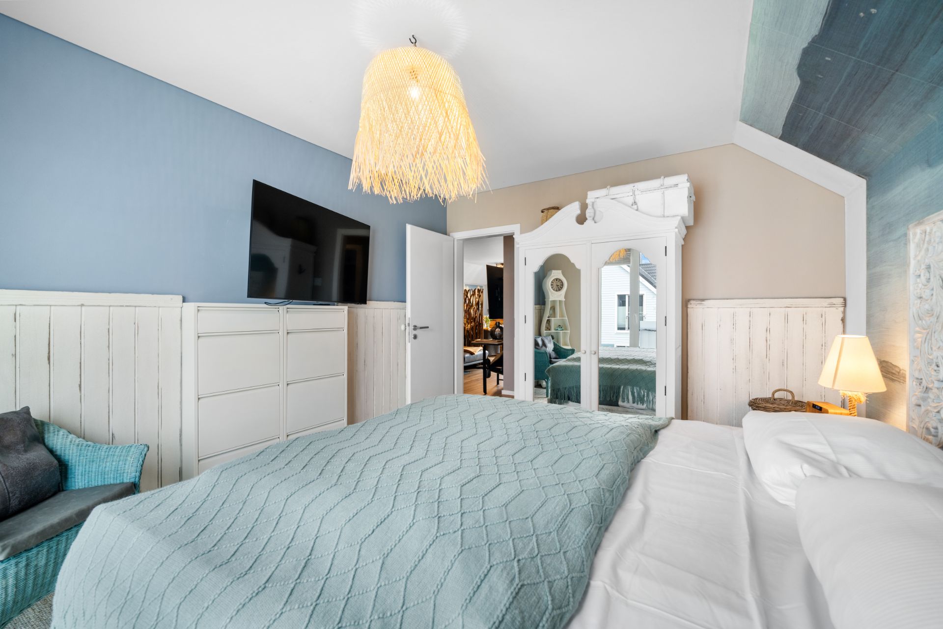 Helles Schlafzimmer mit Doppelbett, TV, Spiegel, blauer Wand und geflochtener Lampe.