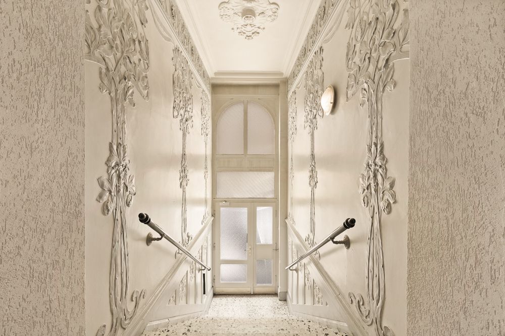 Heller Treppenhausflur mit weißen Stuckverzierungen an den Wänden und einem Glas-Eingangstür.