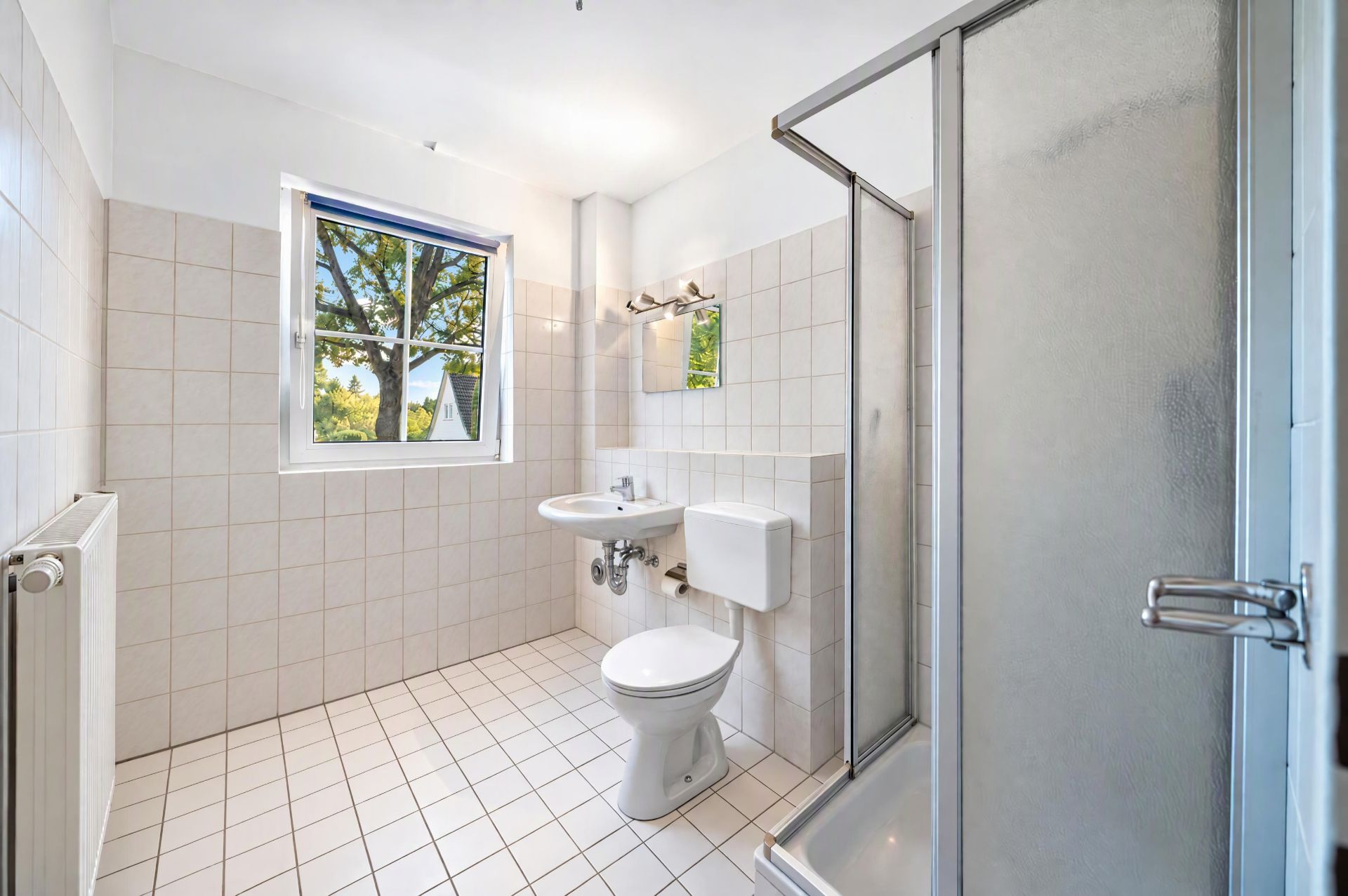Helles Badezimmer mit Fenster, Dusche, Toilette und Waschbecken auf weißen Fliesen.