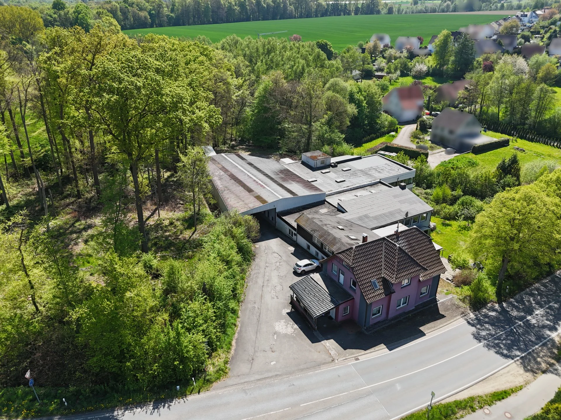 Luftaufnahme eines Wohnhauses mit großem Nebengebäude am Waldrand, umgeben von Bäumen und Feldern.