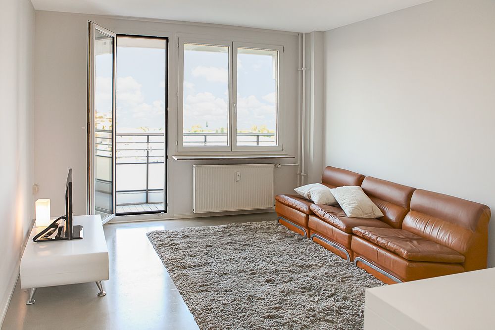 Helles Wohnzimmer mit braunem Ledersofa, großem Teppich und Zugang zu einem Balkon.