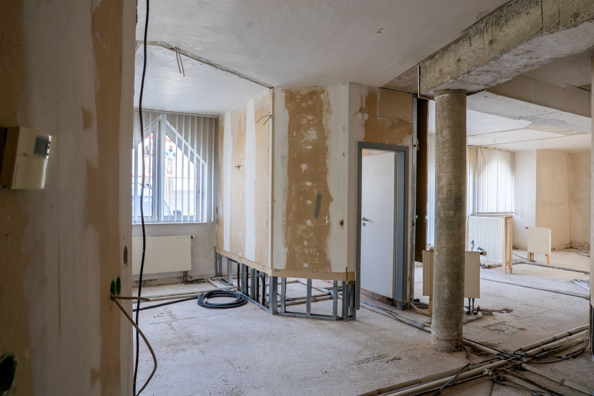 Wohnraum im Umbau mit offenen Wänden, sichtbaren Leitungen und Fenstern mit Jalousien.