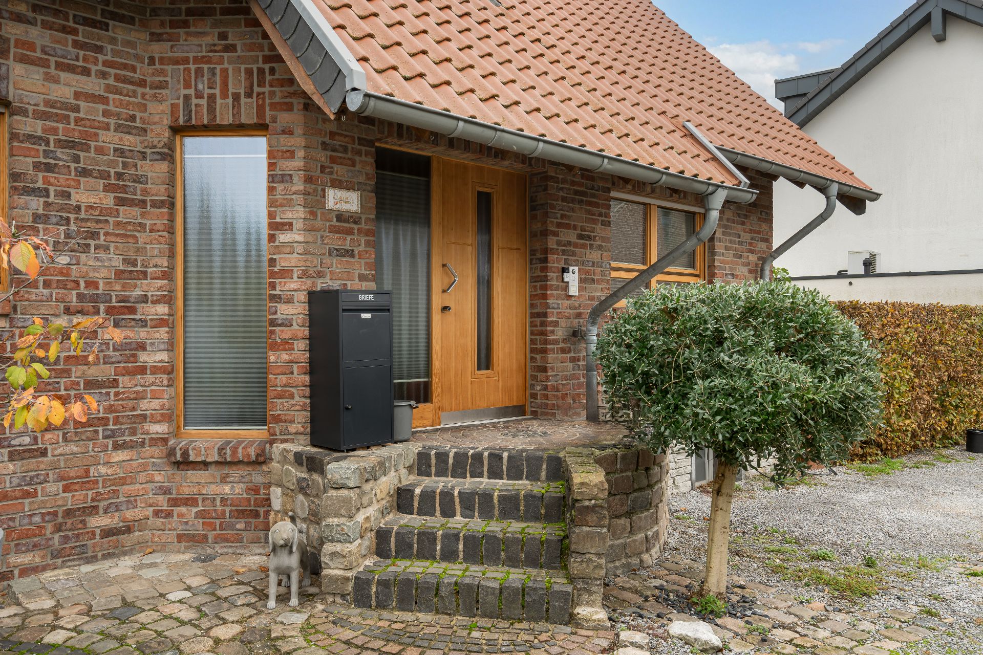 Eingang eines Hauses mit Holztür, Briefkasten, Treppe, kleiner Statue und Strauch.
